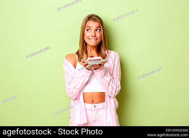 Image of attractive blond girl in pink outfit, looking thoughtful, holding piece of delicious cake, standing over green background
