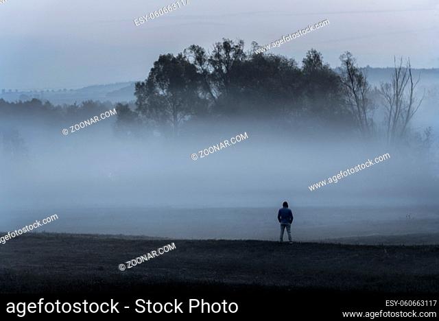 Dramatic nature landscape with a man standing, alone in the cold, on a meadow, contemplating the dense mist, near Schwabisch Hall, Germany