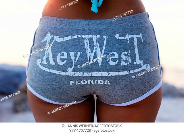 A tan young woman on the beach, seen from behind, wearing hot pants with the writing Key West, Florida across the back