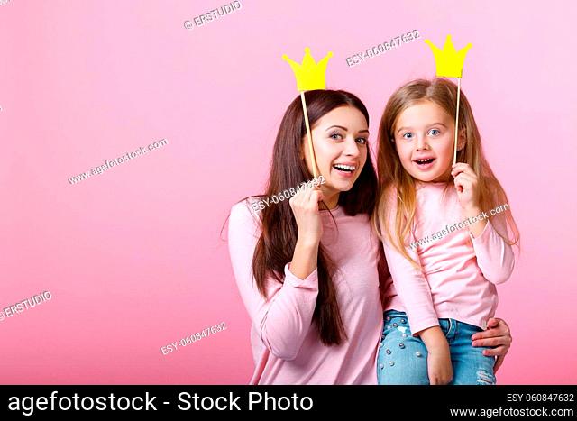 young mother and daughter having fun together and holding paper yellow crown on pink background