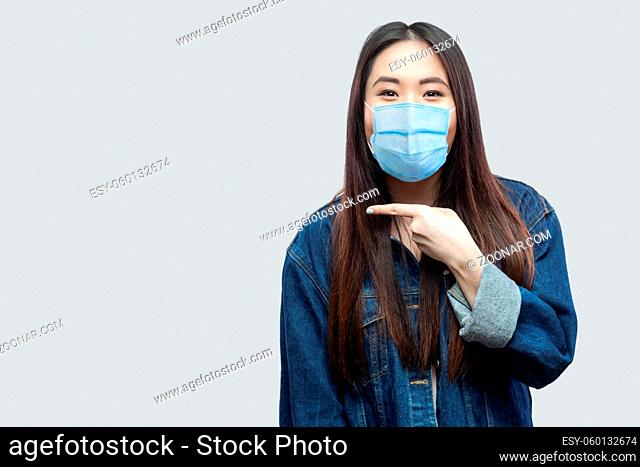 Portrait of excited beautiful brunette asian young woman with medical mask in casual blue denim jacket standing and pointing at copyspace with surprised face