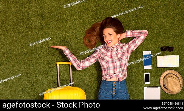 Portrait of beautiful tourist lady with red lips lying on green grass near yellow luggage and many other things: mobile or smart phone, passport, cap