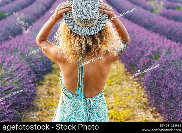 Back view of pretty curly blonde woman holding trendy hair and admiring a scenic lavender field. Travel female people lifestyle