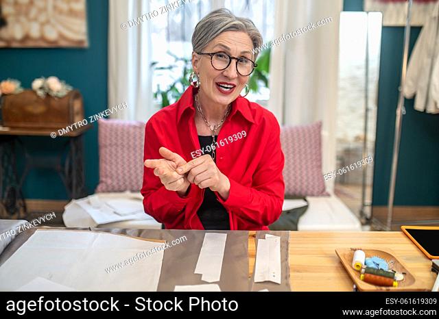 Reasoning. Gray-haired pretty woman in glasses looking at camera convincingly speaking curling fingers sitting at table with pattern in atelier