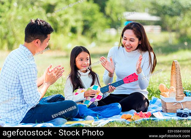 Happy Asian young family father, mother and children having fun and enjoying outdoor together sitting on the grass party with playing Ukulele during a picnic in...