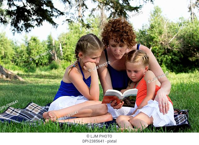 A mother reads a book to her daughters in a park; Stony Plain, Alberta, Canada