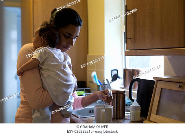 Young mother making a cup of tea with her baby son on her shoulder
