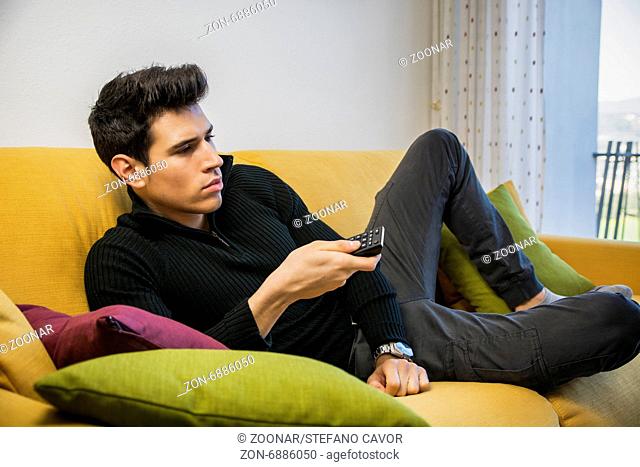 Young man sitting on couch watching television changing the channel with the remote control