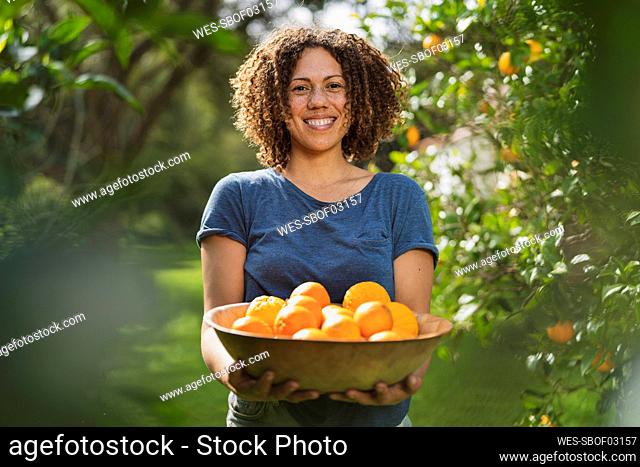 Smiling curly haired woman holding basket of oranges while standing in garden