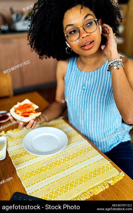 Young woman with curly hair wearing wireless headphones while having breakfast at table