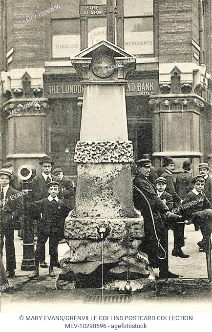 The Aldgate Pump, London. A group of young lads and cheeky street types surround the venerable pump. To the left of the pump is a hand-crank 'Fire Call Post' -...