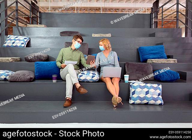 Male and female colleague in face masks sitting in foyer discussing and using tablet and laptop
