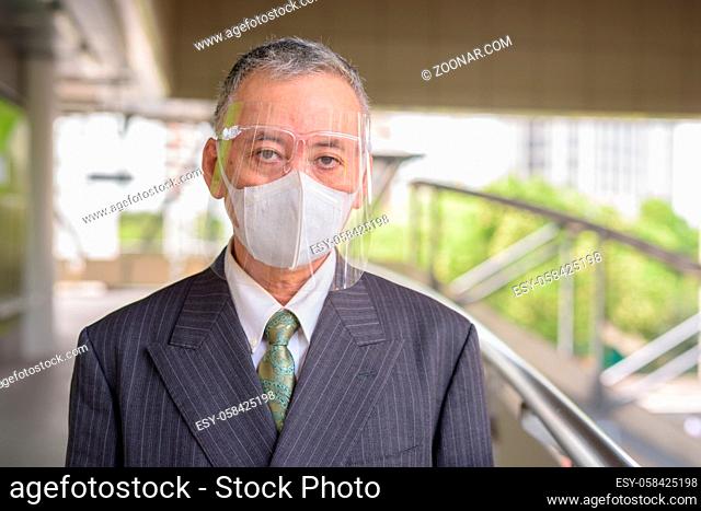 Portrait of mature Japanese businessman with mask and face shield for protection from corona virus outbreak at footbridge