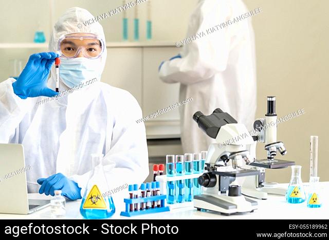 Scientists hold blood sample test tube and examine for his research and develop vaccine for coronavirus covid-19 pandemic with his colleague in background