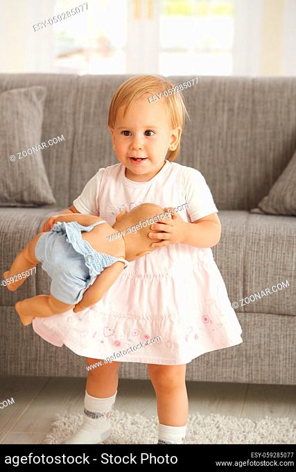 Cute little toddler girl standing in living room holding dolly, smiling