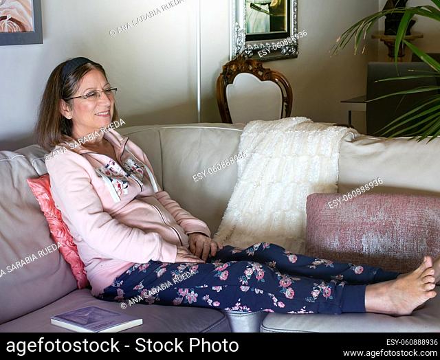 A relaxed older woman enjoys watching TV stretched out on the sofa at home