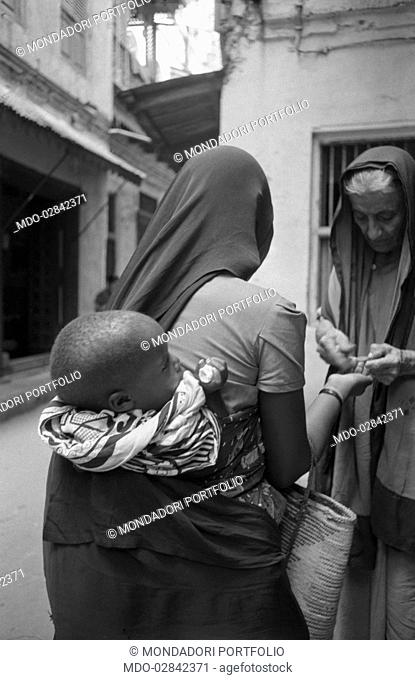Old woman giving something to a woman carrying a baby wrapped on her back. Zanzibar, 1950s