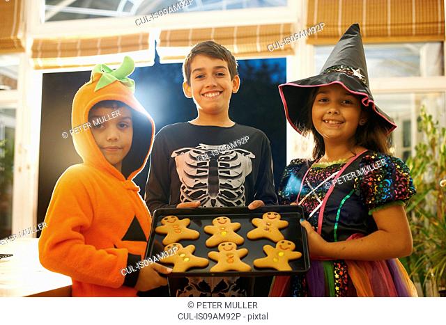Brothers and sister wearing halloween costumes holding tray of gingerbread men