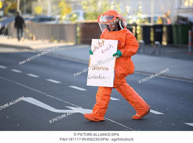 17 November 2018, Hessen, Büdingen: A demonstrator dressed in a protective suit walks across a street during protests against the NPD with a poster saying...