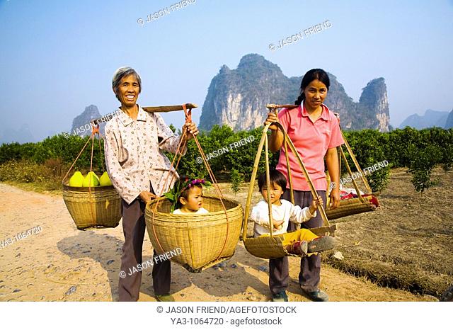 China, Guangxi Zhuang Autonomous Region, Yangshuo County  Two rural farm workers carry fruit and their children in hand woven baskets along a rural road running...