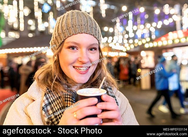 Close-up of happy beautiful woman holding hot chocolate in Christmas market at night
