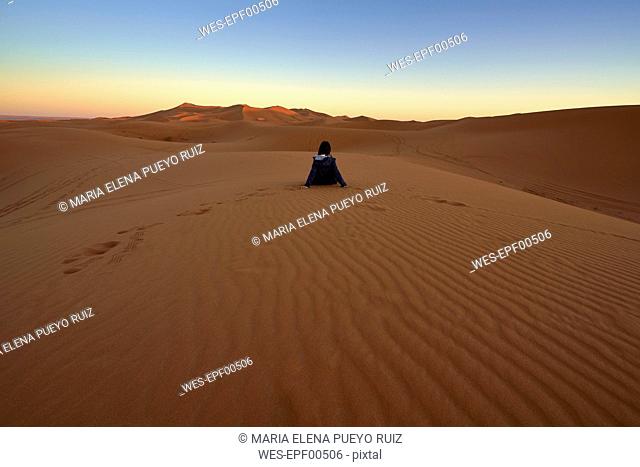 Morocco, back view of woman sitting on desert dune at twilight