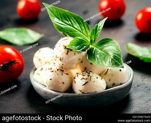 Close up view of small mozzarella cheese marinated with herbs. Mozarella balls with fresh basil and cherry tomatoes on black concrete background