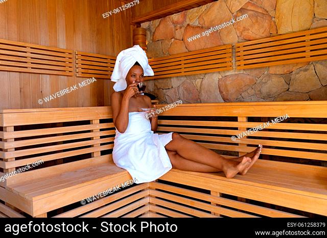 African american young woman wearing bathrobe drinking tea while sitting on wooden bench at sauna
