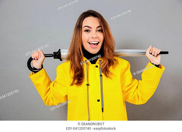 Portrait of a happy girl dressed in raincoat and rubber boots posing while standing with an umbrella and looking at camera isolated over gray background