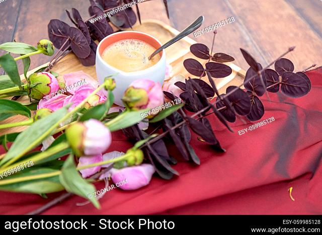 A white Cup of black coffee stands on a wooden background next to pink peonies and other garden flowers and leaves, The concept of village life, Cottagecore
