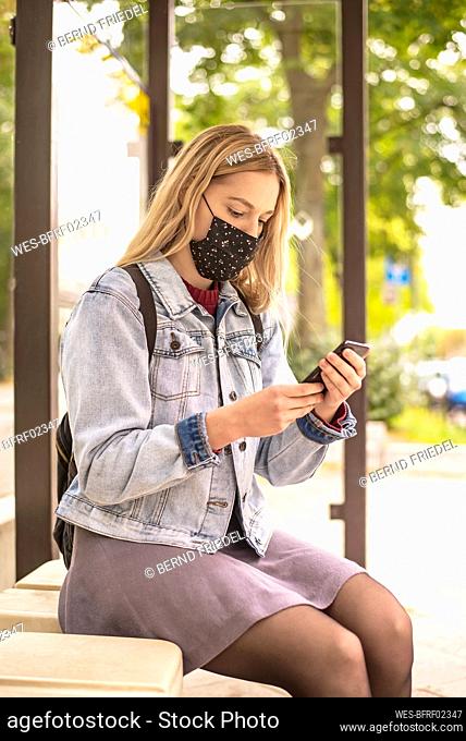 Young woman using smart phone while sitting at bus stop during pandemic