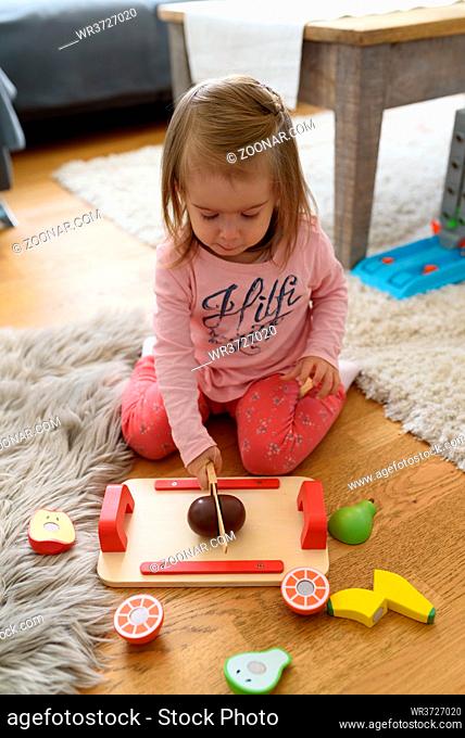 2 year old baby girl at home cutting woden toy fruits with wooden knife. Child development concept