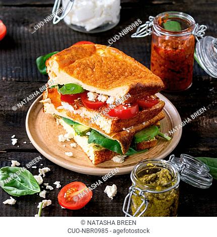 Low-carb gluten free Cloud bread veggie sandwich with spinach, avocado, feta cheese, tomatoes and pesto sauce, served on plate with jars of ingredients over old...