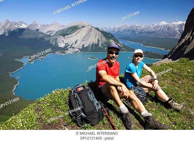 Male and female hiker sitting on top of a mountain ridge overlooking emerald lakes and mountain ranges with blue sky, kananaskis provincial park alberta canada
