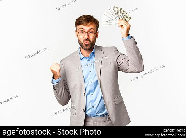 Successful bearded businessman in grey suit and glasses, winning cash, holding money and triumphing, standing happy over white background