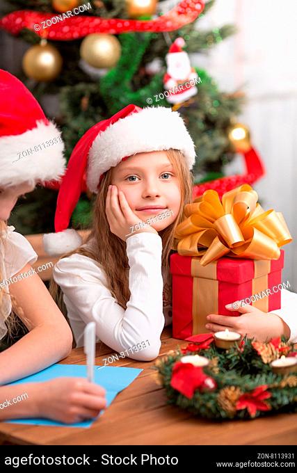 Little girl in Santa hat sitting with present and looking at the camera while her sister writing New Year and Christmas congratulation