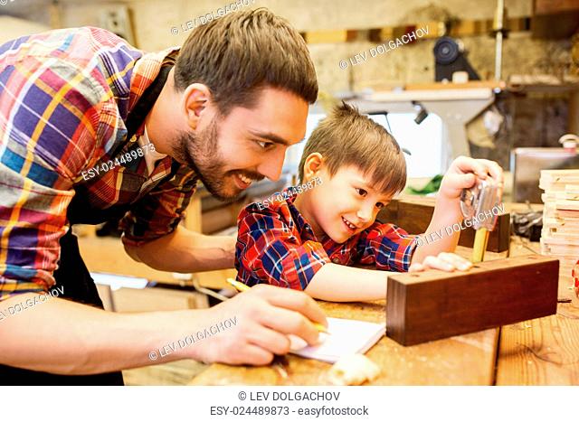family, carpentry, woodwork and people concept - father and little son with ruler and pencil measuring wood plank and writing dimensions to notebook at workshop