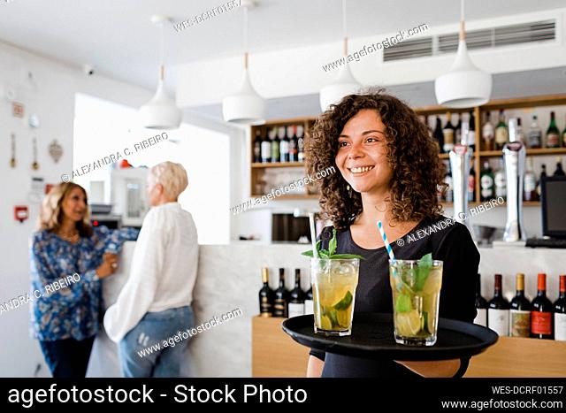Happy young waitress with tray serving cocktail drinks at restaurant