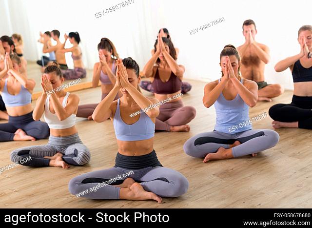 Group of young sporty attractive people in yoga studio, practicing yoga lesson with instructor, sitting on floor in Sukhasana meditative yoga pose