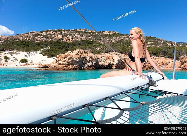 Woman in bikini tanning and relaxing on a summer sailin cruise, sitting on a luxury catamaran near picture perfect white sandy beach on Spargi island in...