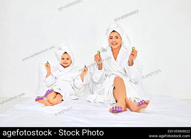 happy woman and little daughter in white bathrobes and towels on heads with pedicure sponge on fingers holding fresh cucumber slices