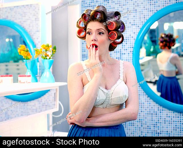 Vintage-styled woman with a surprised look on her face, while wearing hair curlers in a mid-century bathroom