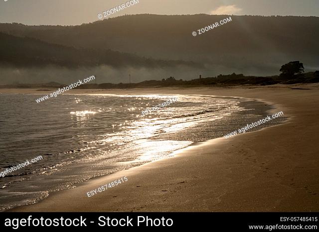 A beautiful curved golden sandy beach with glistening waves in the morning light and fog in the background hills