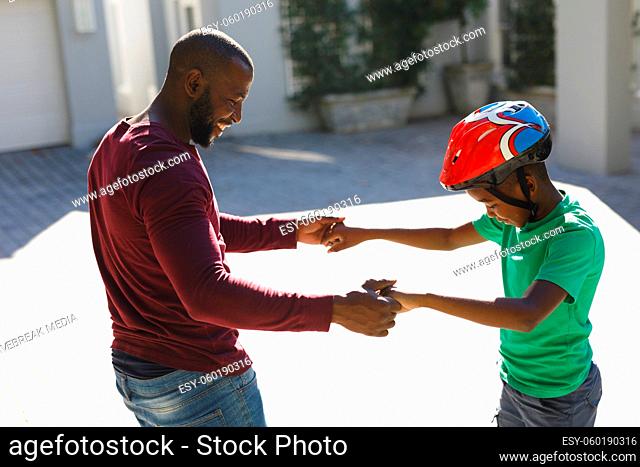 African american father smiling and helping son balancing on skateboard in garden