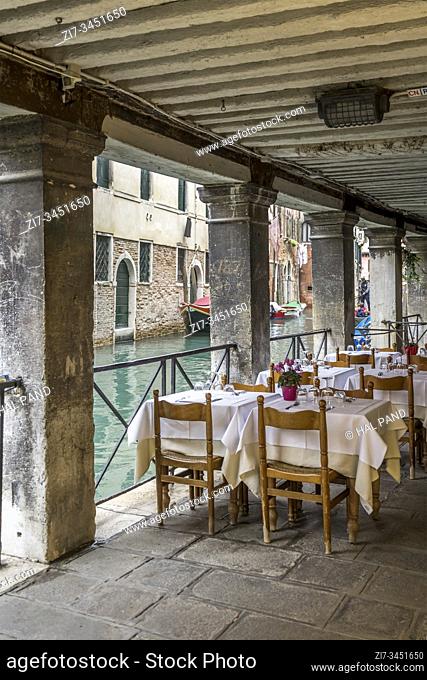 VENICE, ITALY - October 30 2019: cityscape with empty restaurant furnished tables at covered walkway under historical houses on canal