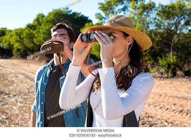 Young woman looking through binoculars while boyfriend drinking water in forest