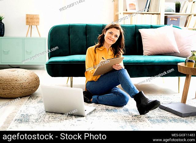 Smiling mature woman writing on clipboard while sitting at sofa in living room