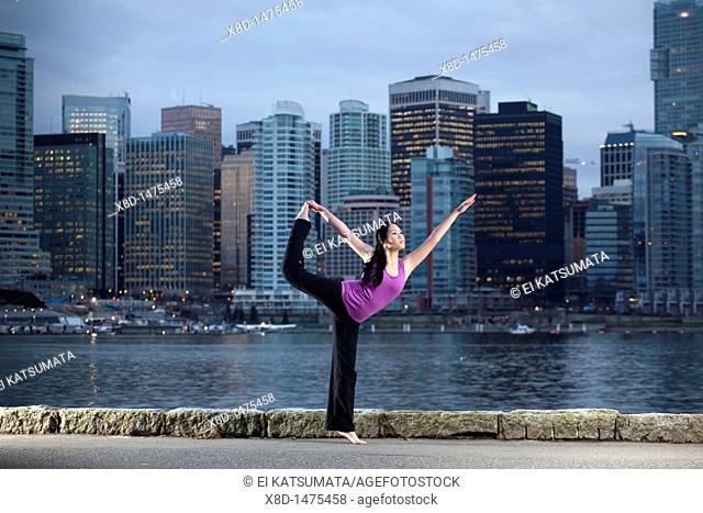 A young Asian woman does the Natarajasana Lord of the Dance yoga pose along the Stanley Park seawall with the city skyline beyond, Vancouver, British Columbia