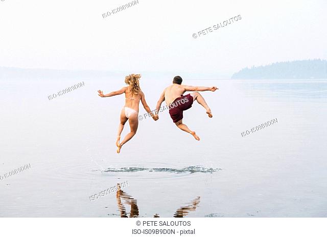 Young couple on beach, holding hands, jumping, mid air, rear view