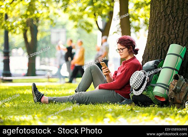 Girl teenager tourist with backpack looking at smartphone in city park, telephoto shot